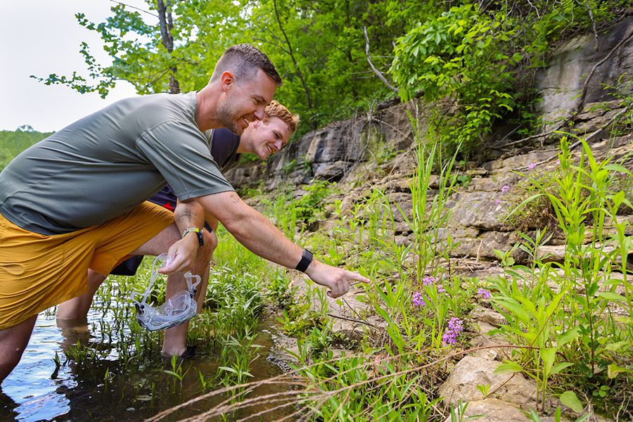 Educator and guest standing in the creek pointing at a something in the tall grass.