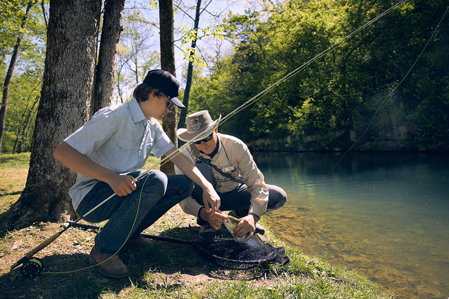 A man holds a fly fishing rod while another man offers instructions on how to attach the bait.